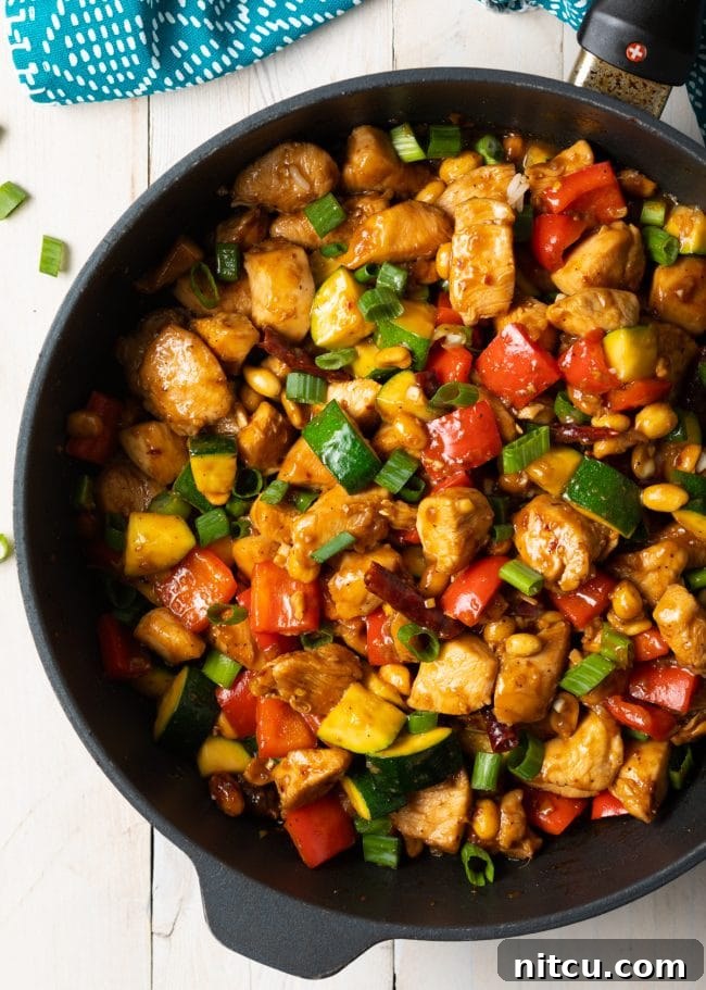 Sichuan Kung Pao Chicken 10 Top down view of a cast iron skillet with chopped cooking chicken, peanuts, chopped scallions, zucchini and bell pepper.