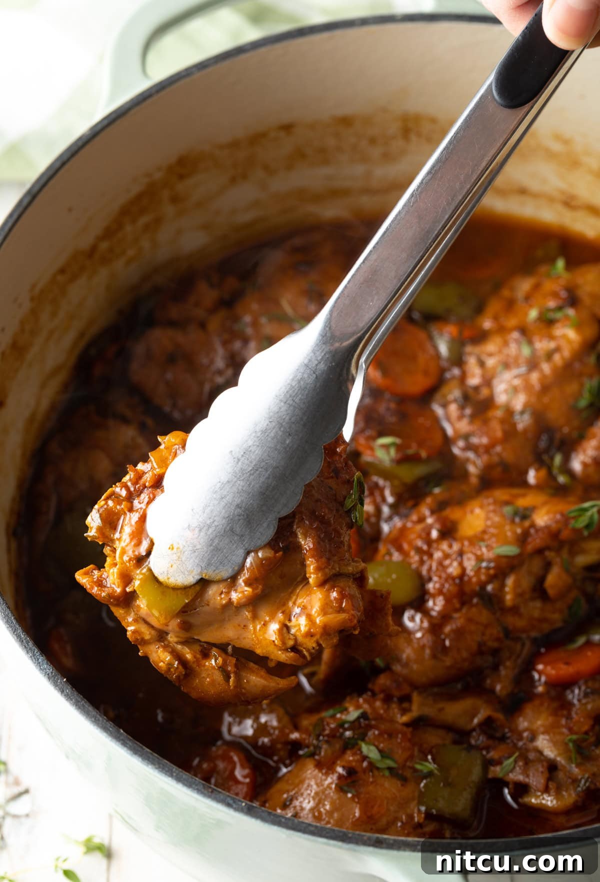 Authentic Jamaican Brown Stew Chicken 7 Pot filled with ready to serve stew and a tong removing a piece of chicken.