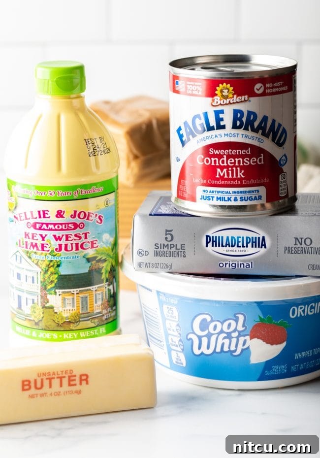Effortless Key Lime Pie 5 A collection of key ingredients laid out: softened cream cheese, a tub of Cool Whip, bottled key lime juice, sweetened condensed milk, sticks of butter, and a stack of graham crackers, ready for pie making.
