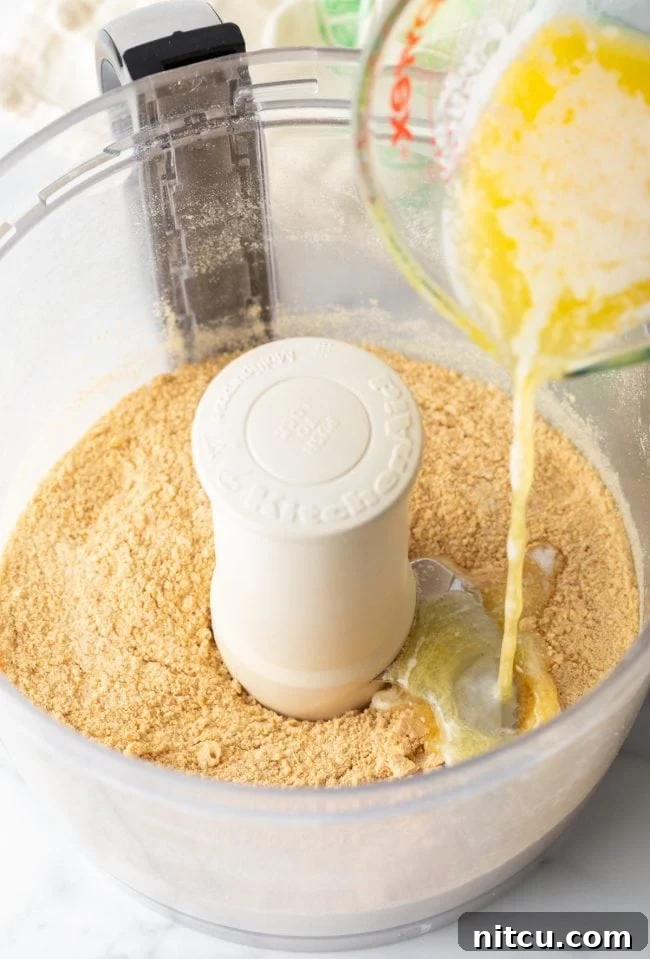 Effortless Key Lime Pie 6 Graham cracker crumbs inside a food processor bowl, with a stream of melted butter being poured over them, ready for blending to form the pie crust.