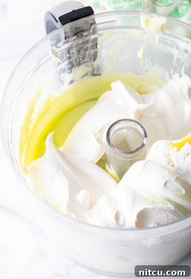 Effortless Key Lime Pie 9 A large scoop of fluffy Cool Whip being added to the bright key lime filling mixture inside a food processor bowl, preparing for the gentle folding process.