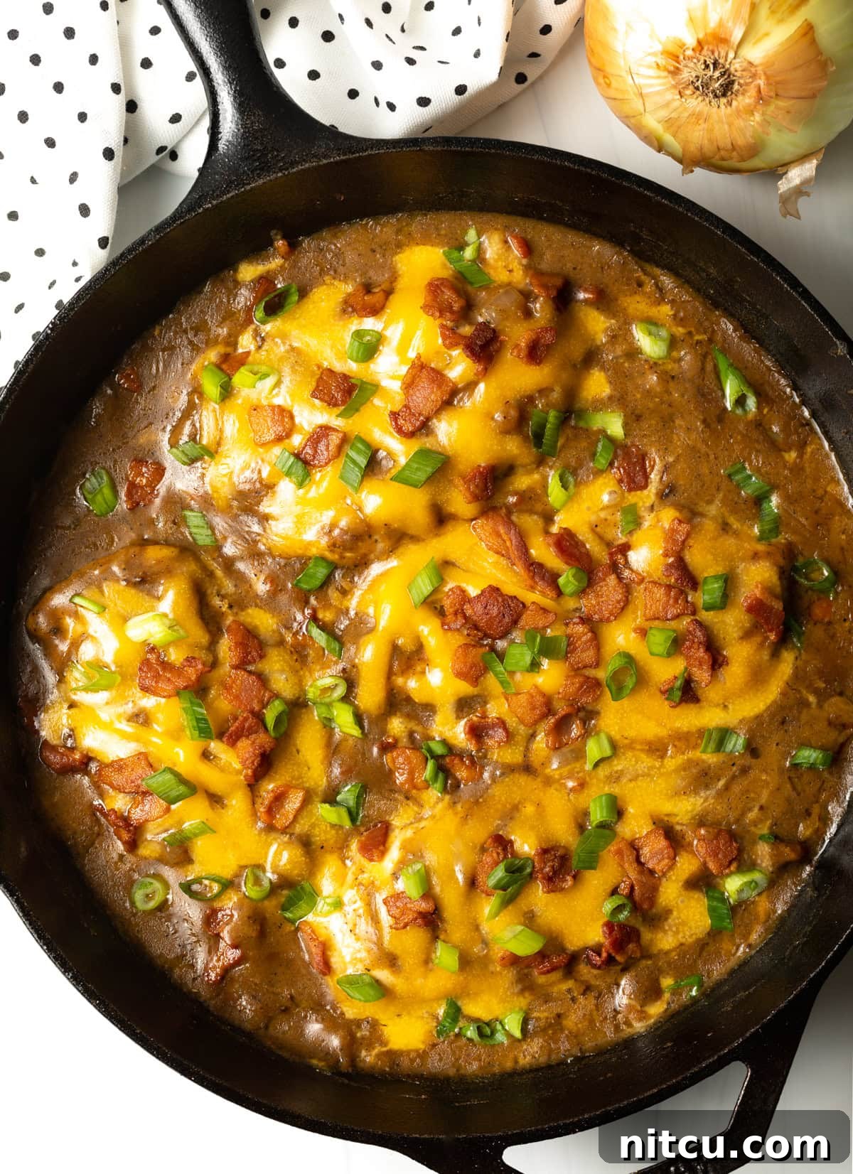Braised Chicken Delight 3 Overhead view of a cast iron skillet filled with golden-brown chicken, rich gravy, melted cheese, crispy bacon, and fresh green onions.