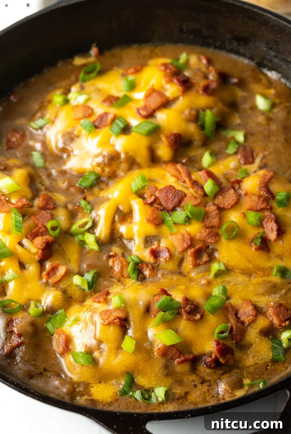 Braised Chicken Delight 10 Close up side view of smothered chicken and gravy recipe made with cheese, bacon and scallions in a cast iron skillet, ready to be served.