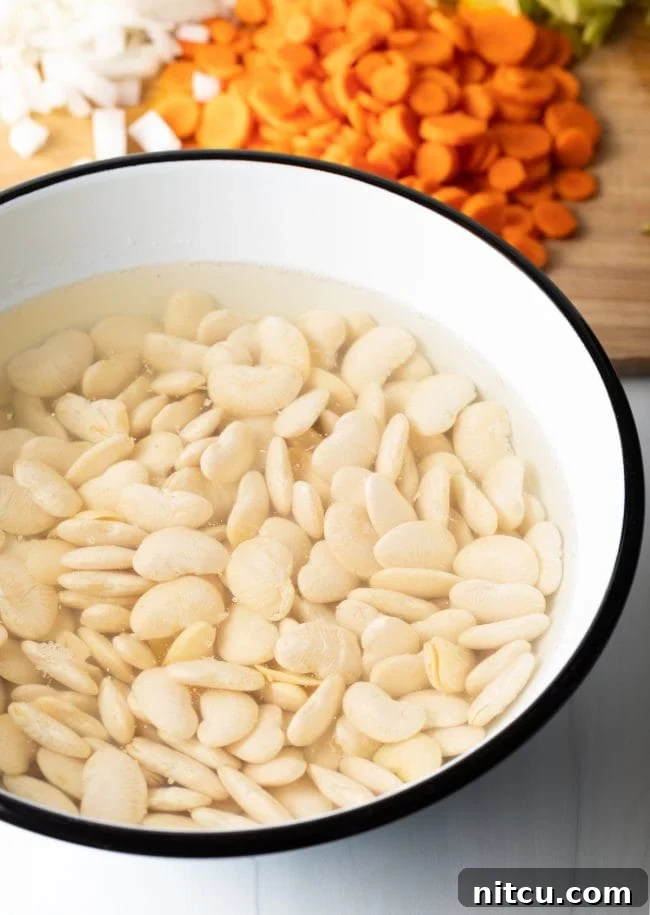 Dried butter beans soaking in a clear bowl of cold water on a countertop, indicating the first step of preparation.