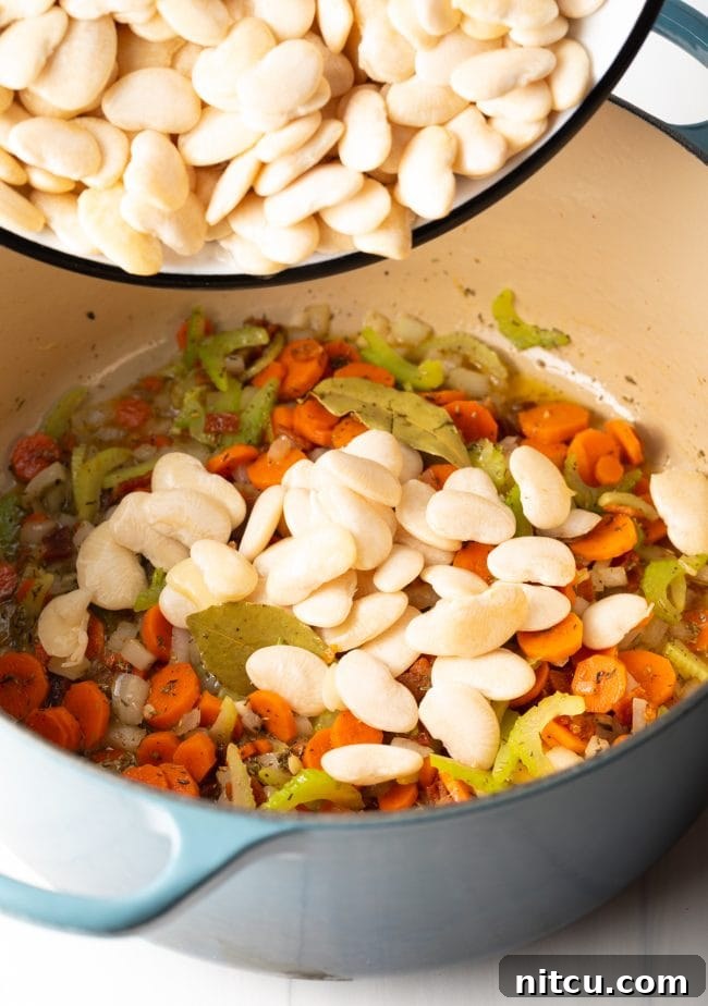 Perfectly soaked lima beans being carefully added to a large pot containing sautéed vegetables and crispy bacon, ready for the next cooking stage.