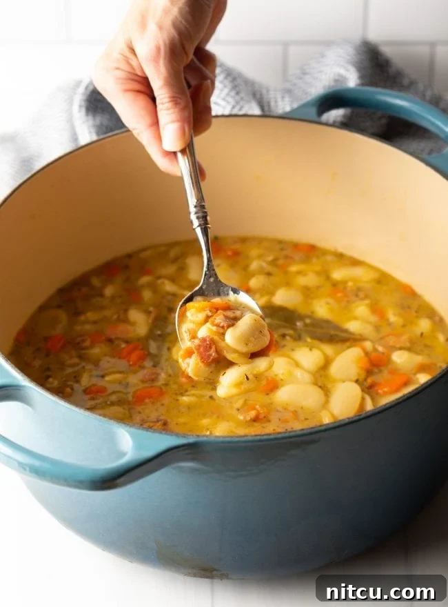 A hand using a large serving spoon to scoop steaming, tender butter beans with crispy bacon bits from a deep pot, ready to be served.