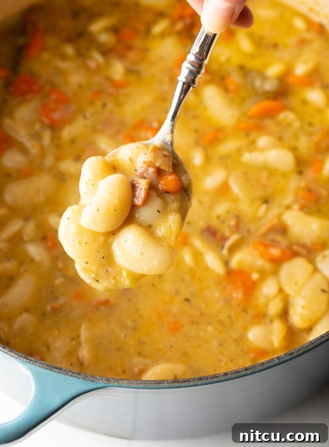 A serving spoon diligently scooping rich, tender butter beans, intermingled with savory bacon, sweet carrots, and fragrant aromatics, from a large, steaming pot.