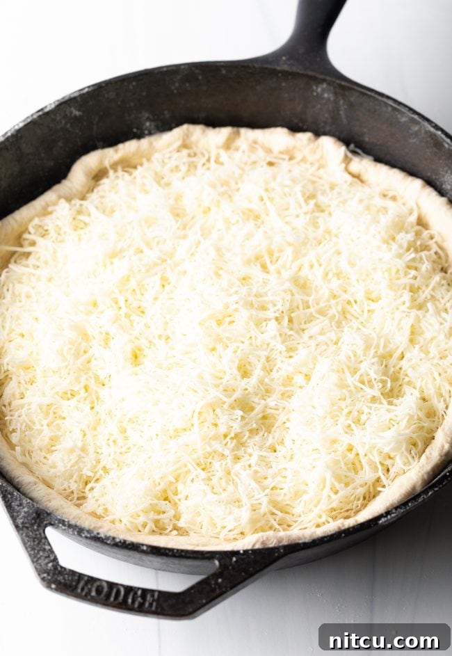 Iron-Clad Deep Dish Delight 5 Shredded Italian cheese blend being meticulously spread across the raw pizza dough within a cast iron skillet, forming the essential first layer of the deep dish pizza.