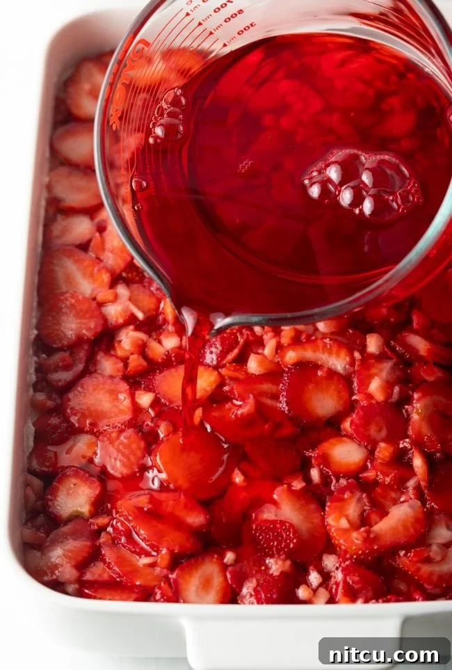 Sweet Strawberry Pretzel Delight 11 Pouring the cooled red jello mixture over the layers of diced strawberries and cream cheese in the strawberry pretzel dessert recipe.