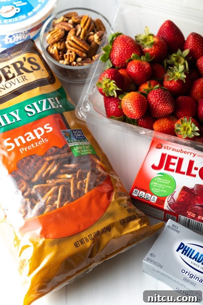Sweet Strawberry Pretzel Delight 4 A top-down view of ingredients laid out: a basket of fresh strawberries, a box of strawberry jello, cream cheese, a bag of pretzels, a bowl of pecans, and a tub of cool whip.