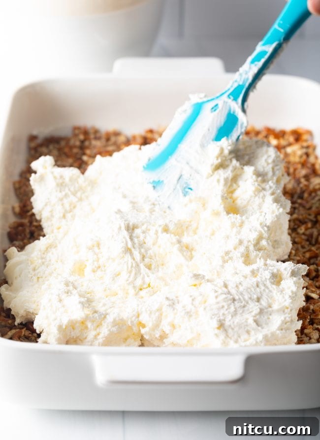 Sweet Strawberry Pretzel Delight 10 A blue spatula spreading a smooth layer of cream cheese and cool whip fluff over the baked pretzel crust in a baking dish.