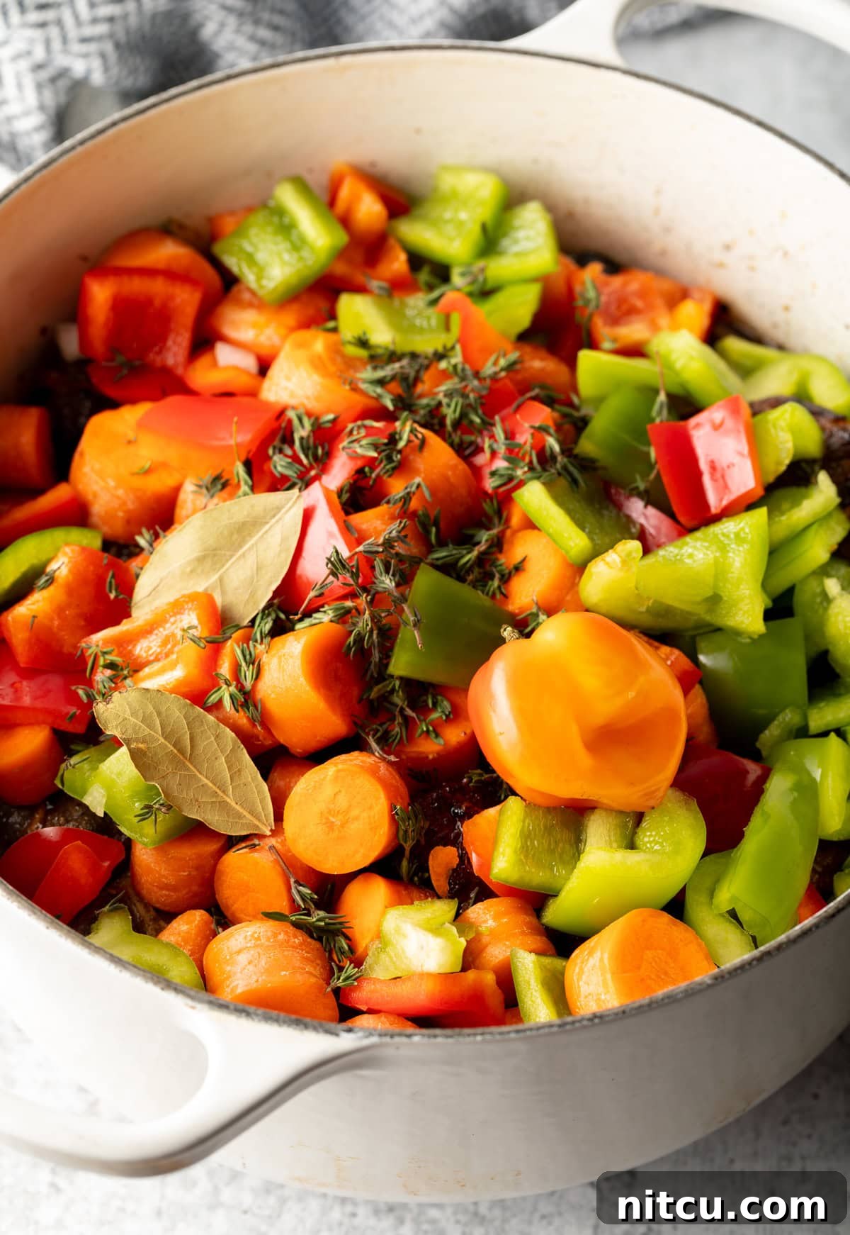 Cooking pot filled with chopped raw vegetables like carrots and bell peppers, along with seasonings, layered over sliced raw oxtail.