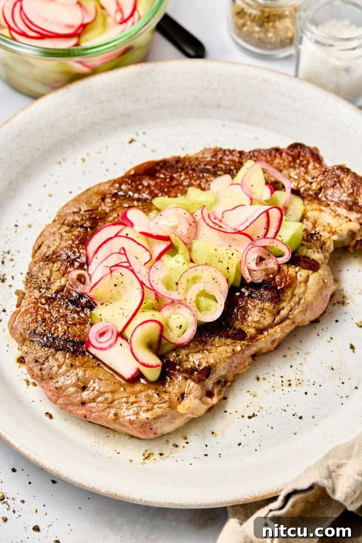 Large grilled steak on a white plate topped with pickled red onions, cucumbers, and radishes.