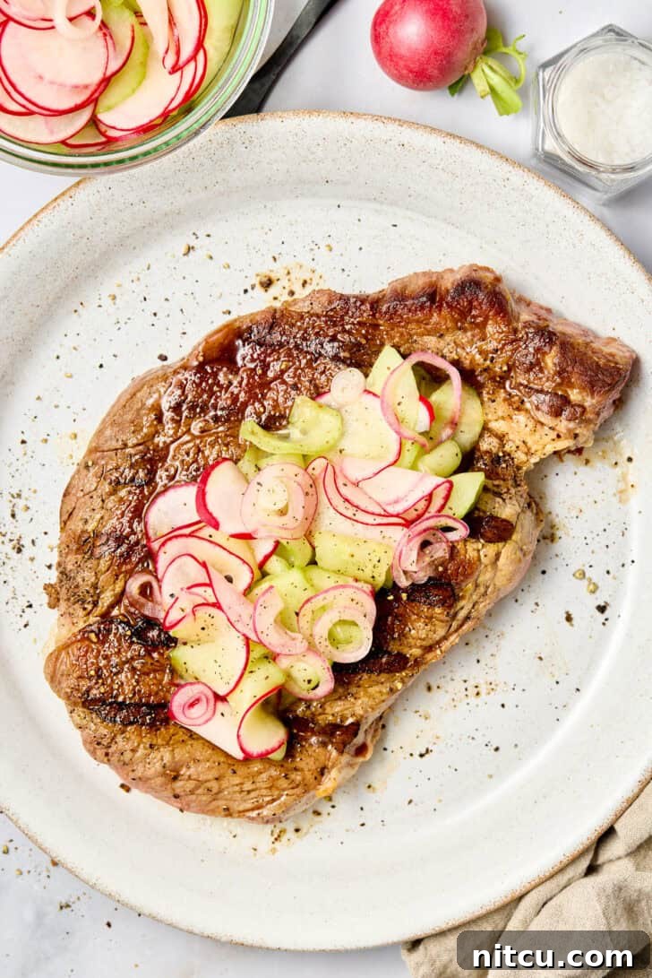 Top down view of a large grilled steak on a white plate topped with pickled red onions, cucumbers, and radishes.