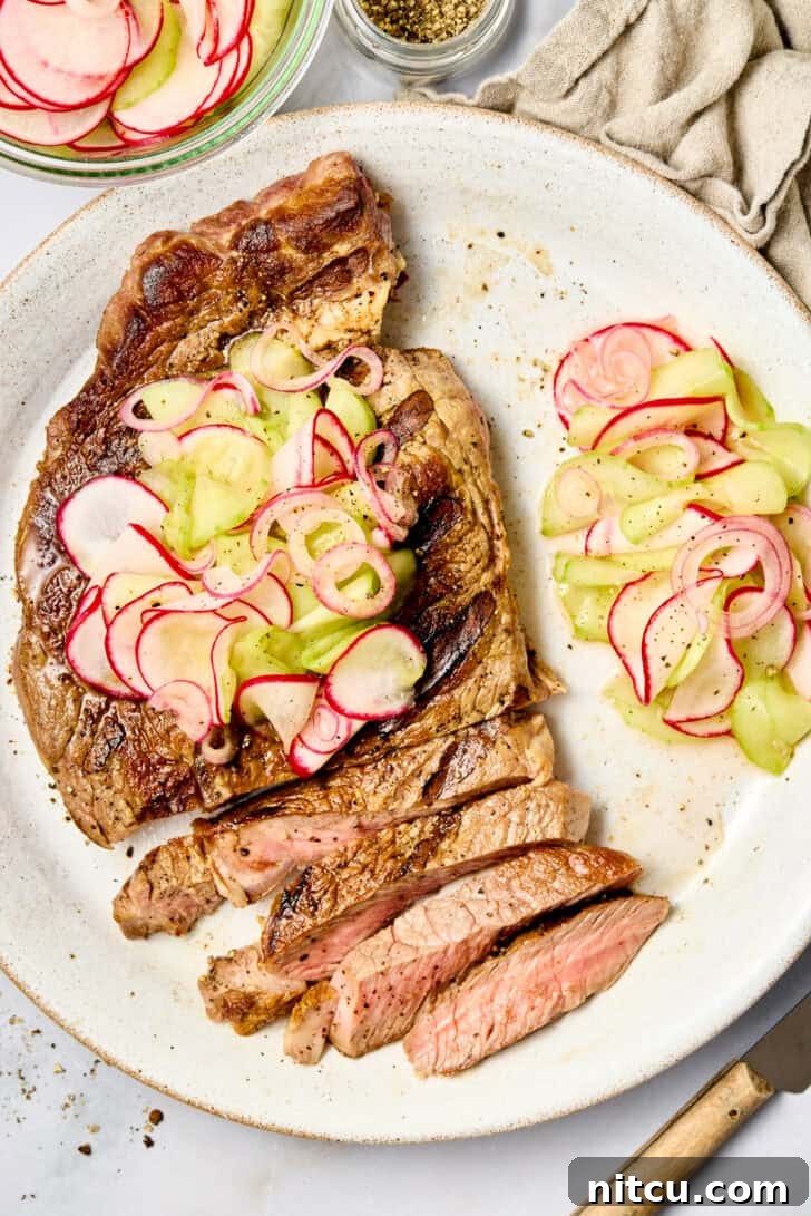 Top down view of a large grilled steak cut into slices on a white plate. The steak has a pickled veggies topping and pile of the veggies on the plate next to the steak.