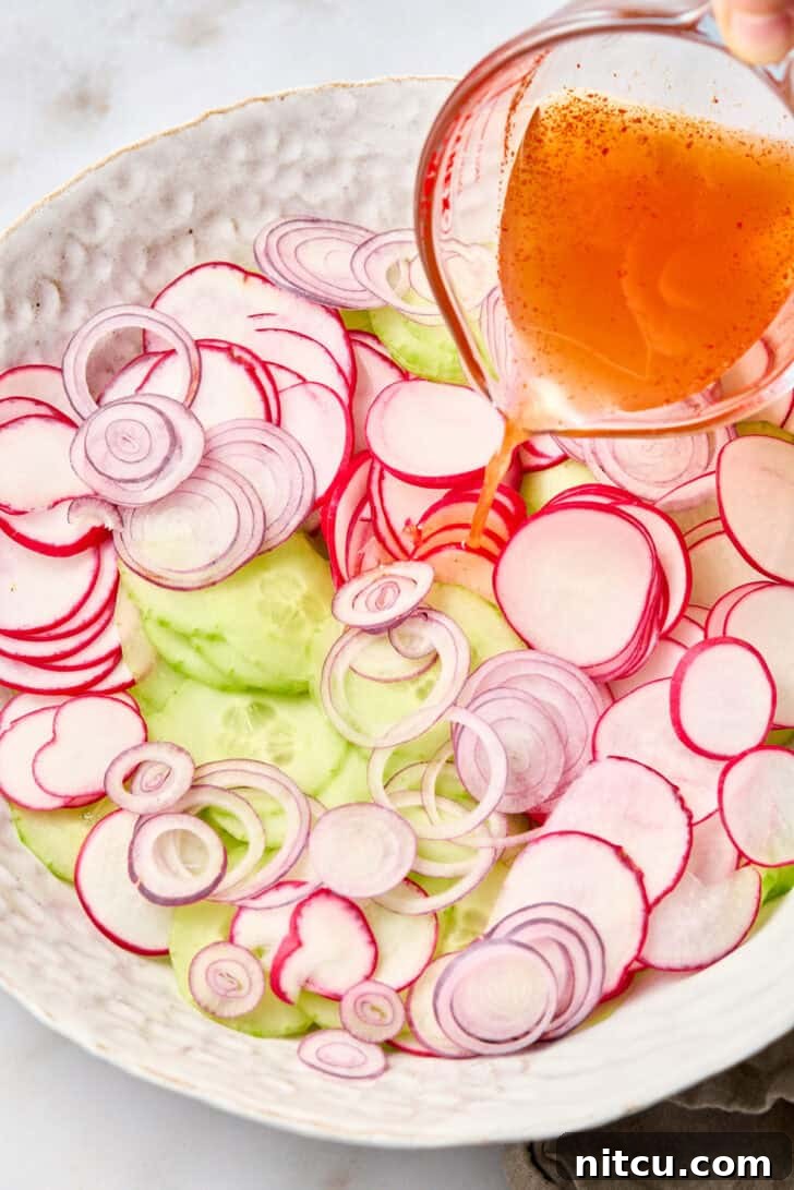 Sliced radishes, cucumbers, and red onion in a large bowl. A hand is pouring a pickling liquid from a glass measuring cup over the veggies.