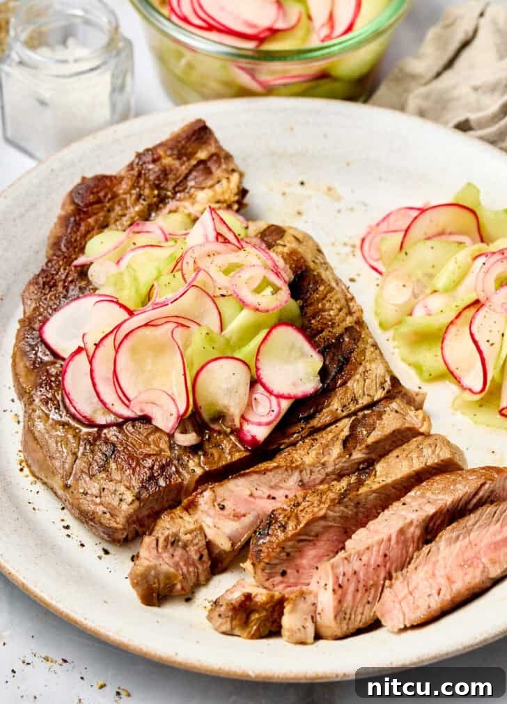 Large grilled steak cut into slices on a white plate. The steak has a pickled veggies topping and pile of the veggies on the plate next to the steak.