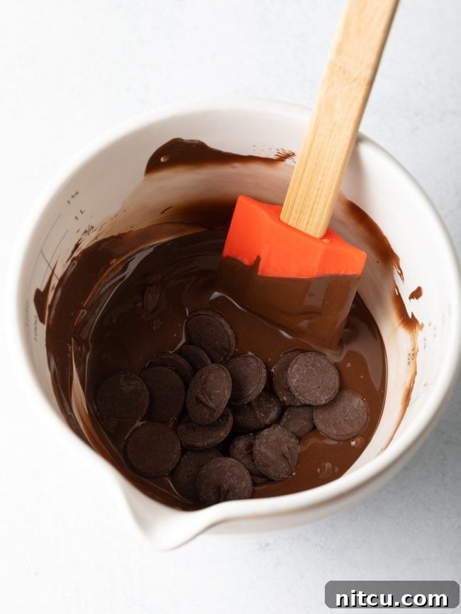 Crackable Chocolate Treasure 6 Glossy chocolate wafers are captured in the process of melting within a white measuring cup, stirred by a clean spatula, ready for molding.