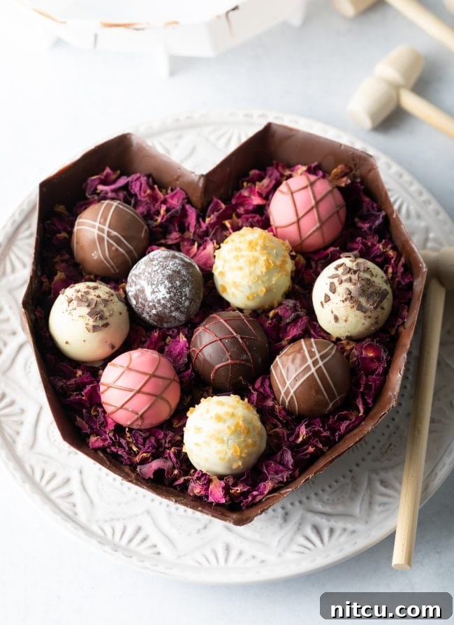 Crackable Chocolate Treasure 10 The bottom half of a breakable chocolate heart rests on a white plate, elegantly filled with delicate edible rose petals and an assortment of gourmet truffles.