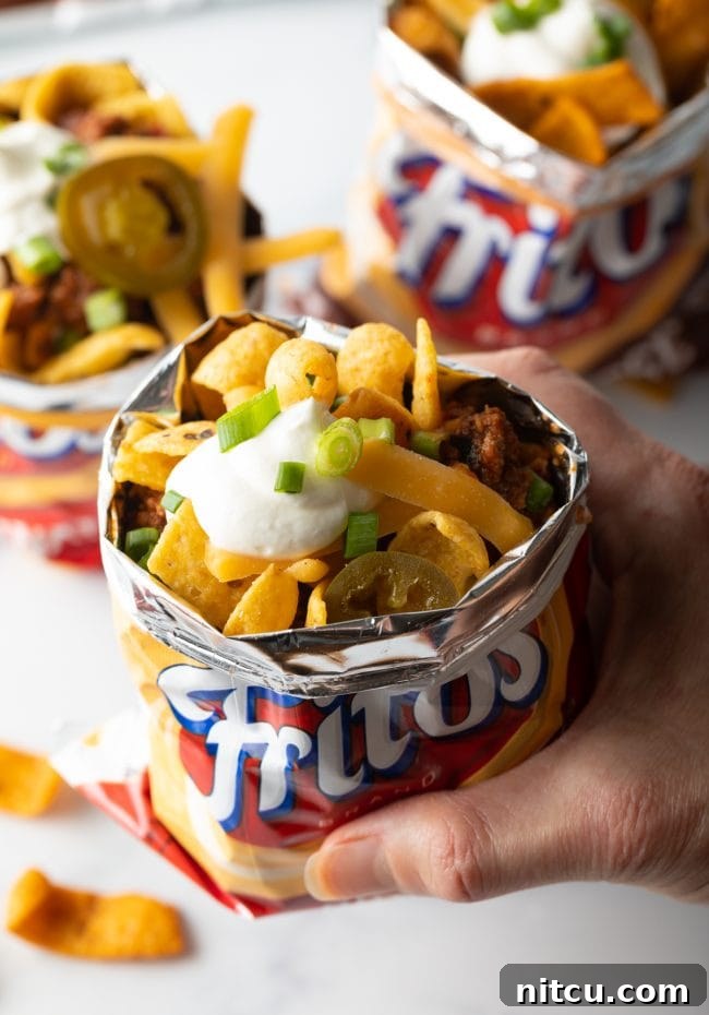 Ultimate Frito Chili Pie 12 A hand holding an open bag of Fritos corn chips filled with a complete Frito chili pie, topped with chili, cheese, and other garnishes.