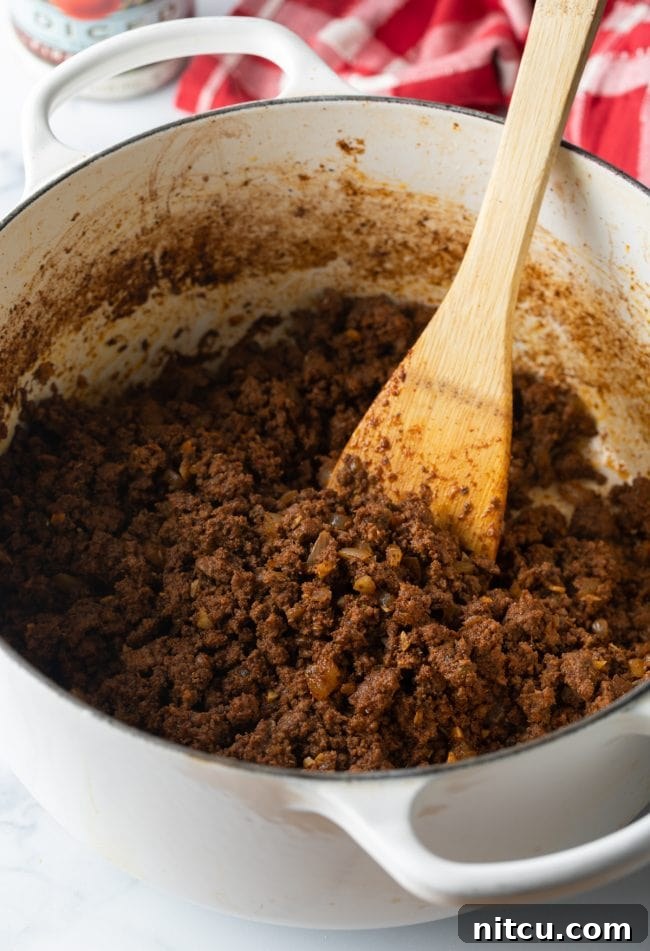 Ultimate Frito Chili Pie 5 Lean ground beef being browned in a large pot, stirred with a wooden spoon, alongside diced onions and garlic.