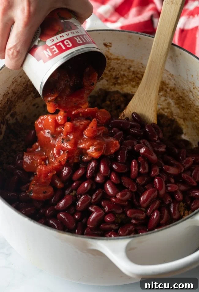 Ultimate Frito Chili Pie 6 A hand adding canned diced tomatoes to a pot containing kidney beans and browned beef, stirred with a wooden spoon.