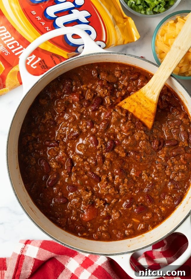 Ultimate Frito Chili Pie 7 A top-down view of a finished pot of rich, simmering chili with a wooden spoon resting inside, ready for serving.