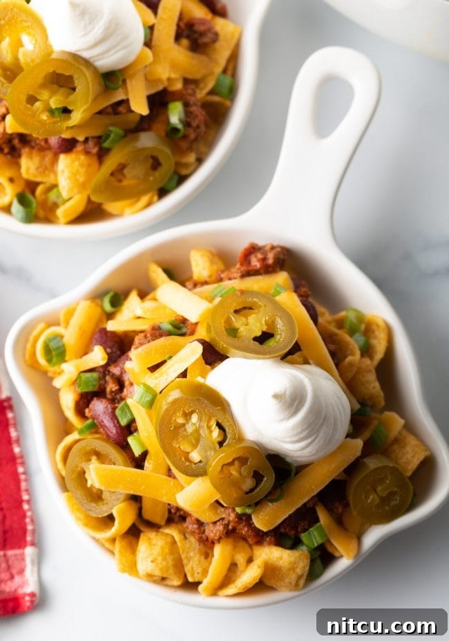 Ultimate Frito Chili Pie 9 A top-down view of a finished Frito chili pie in a shallow white dish, adorned with chili, cheese, scallions, jalapenos, and sour cream.