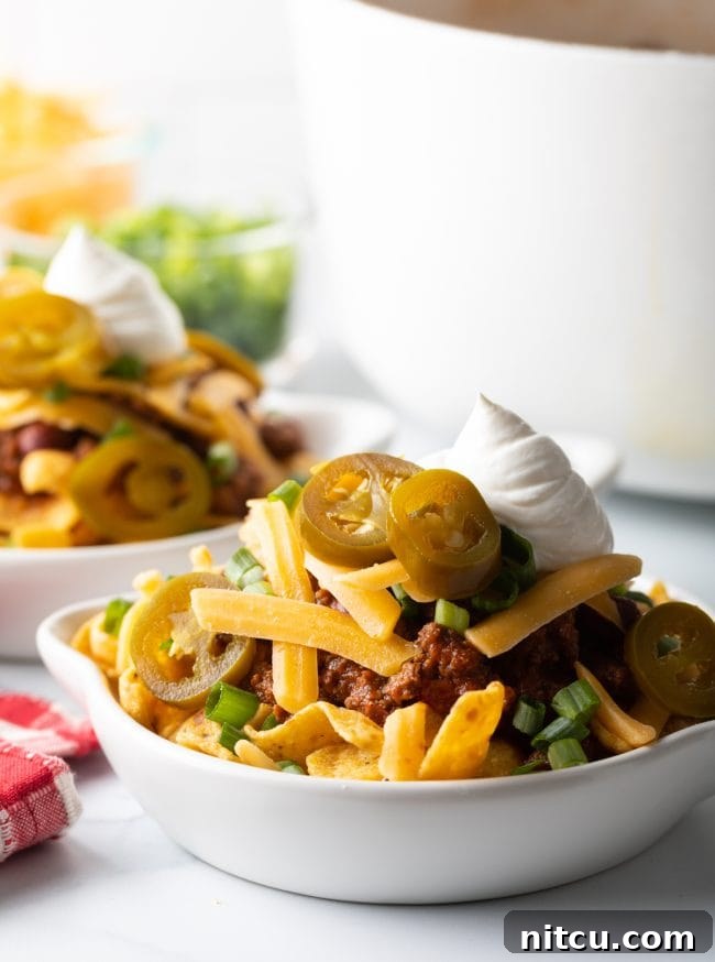 Ultimate Frito Chili Pie 10 A side view of a white shallow dish brimming with Frito chili pie, showcasing layers of beef chili, crunchy corn chips, shredded cheese, chopped scallions, tangy jalapeno slices, and a creamy dollop of sour cream.