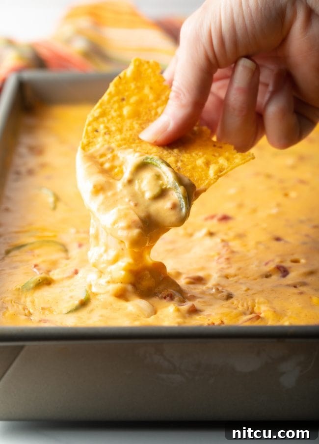 Pitmaster's Smoked Queso 2 Hand scooping creamy smoked queso dip with a jalapeño garnish from a large metal pan, ready for serving.