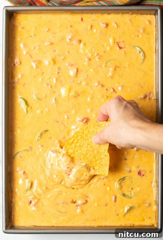 Pitmaster's Smoked Queso 3 Top view of smoked queso dip in a metal pan, a hand is dipping a tortilla chip into the cheesy goodness.