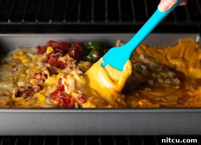 Pitmaster's Smoked Queso 6 A metal pan of smoked queso dip being stirred with a spatula inside a smoker, creating a creamy texture.