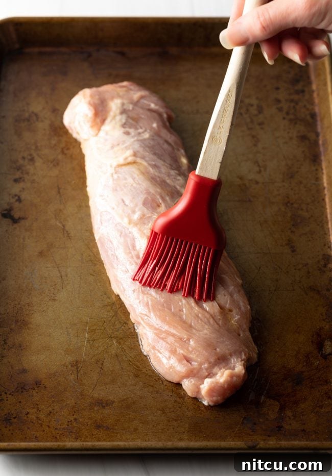 A basting brush applying olive oil to a raw pork tenderloin laid out on a baking sheet, preparing it for seasoning.
