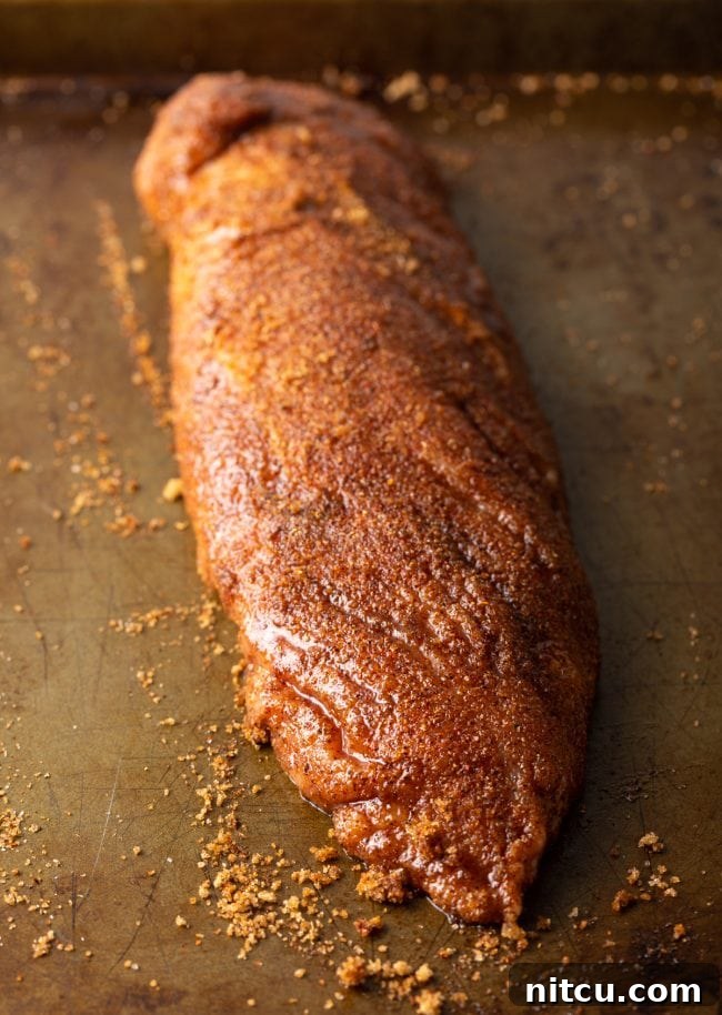 A raw pork loin, visibly covered in a rich, dark spice rub, resting on a baking sheet, ready for the air fryer.