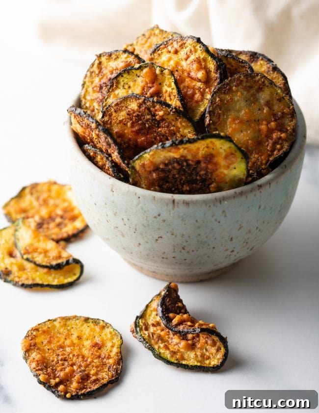 Crispy Air-Fried Zucchini Bites 3 Full view of a speckled bowl overflowing with parmesan zucchini crisps and chips scattered around on a wooden surface.