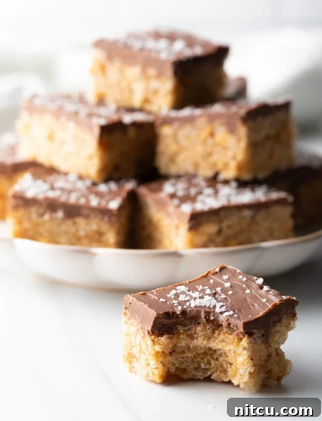 Peanut Butter Rice Krispie Treats 4 Pulled back view showing Scotcharoo with bite taken out in the foreground, plate stacked with squares in the background.