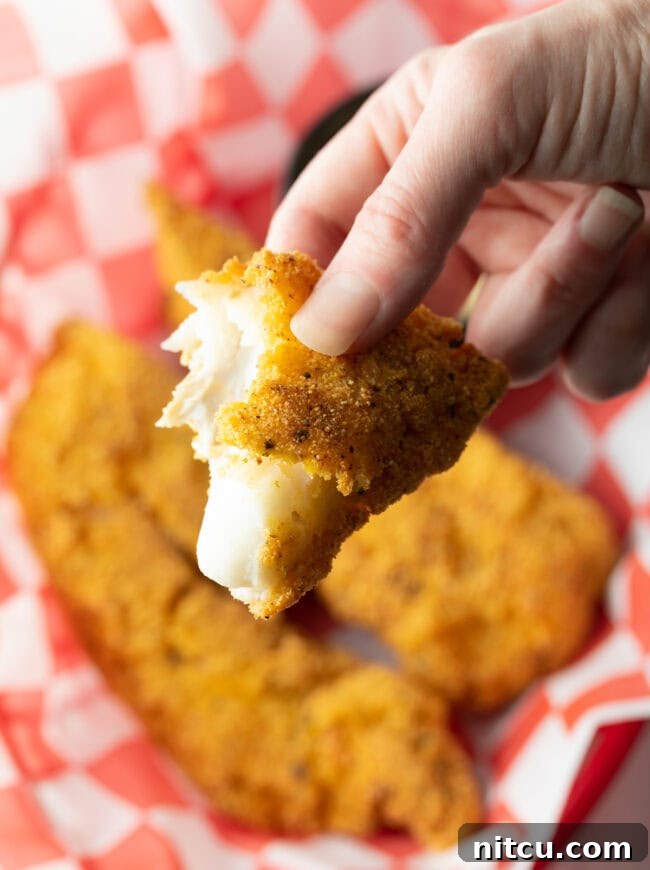 Crispy Corn-Crusted Fish 11 A hand presenting a piece of perfectly fried cornmeal fish towards the camera, showcasing its golden, crispy crust.