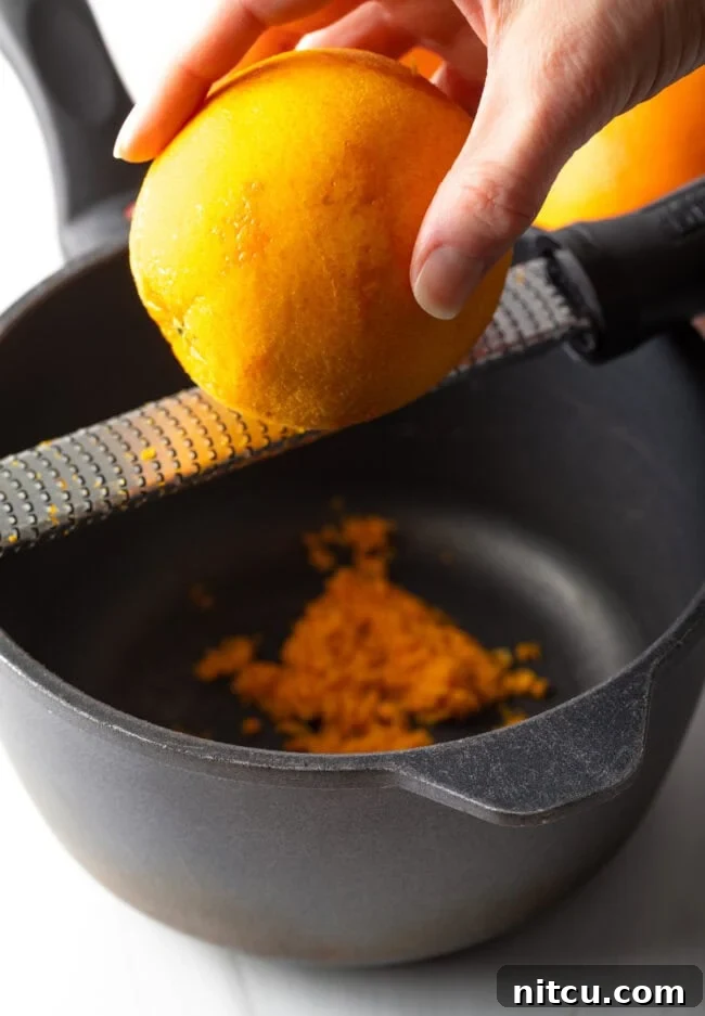 Sunshine Orange Curd 5 Hand zesting an orange above a bowl. A hand skillfully zesting a fresh orange directly over a bowl, capturing the fine, aromatic zest. This image emphasizes the importance of fresh citrus zest in the recipe and the technique for gathering it.