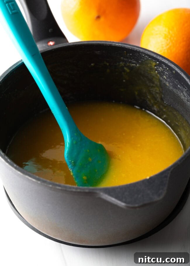 Sunshine Orange Curd 8 Pot cooking orange curd with blue rubber spatula stirring. A blue rubber spatula actively stirring vibrant orange curd in a pot as it cooks on the stovetop. This action shot emphasizes the importance of continuous stirring to achieve a smooth, lump-free curd, preventing it from sticking or burning.