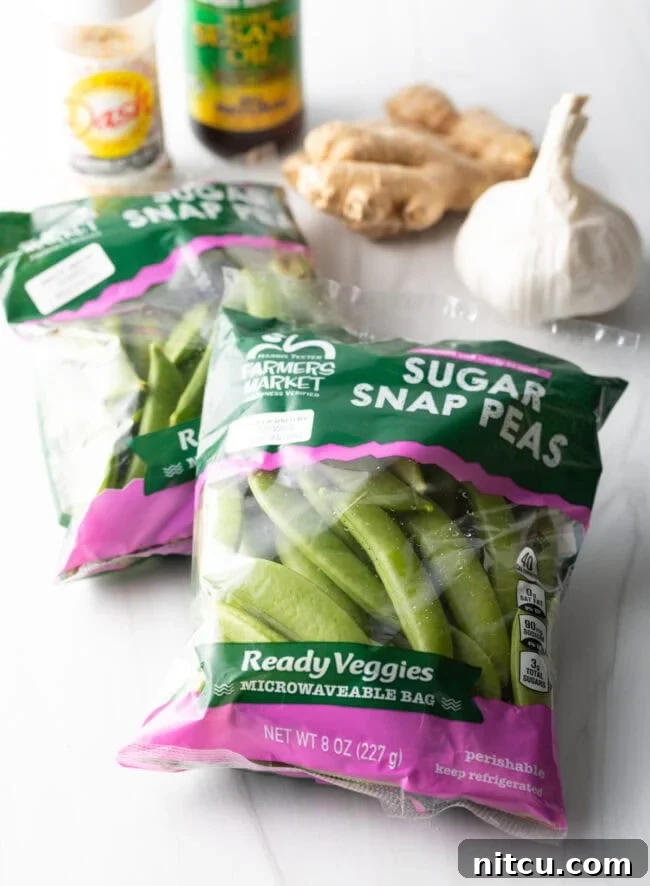 Crispy Garlic Snap Pea Stir Fry 4 Fresh ingredients laid out: a bag of sugar snap peas, a head of garlic, a piece of ginger, and small bowls of seasonings in the background.