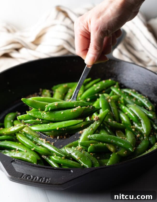 Crispy Garlic Snap Pea Stir Fry 5 A hand stirring bright green sugar snap peas in a large metal skillet with a metal spoon, steam rising.