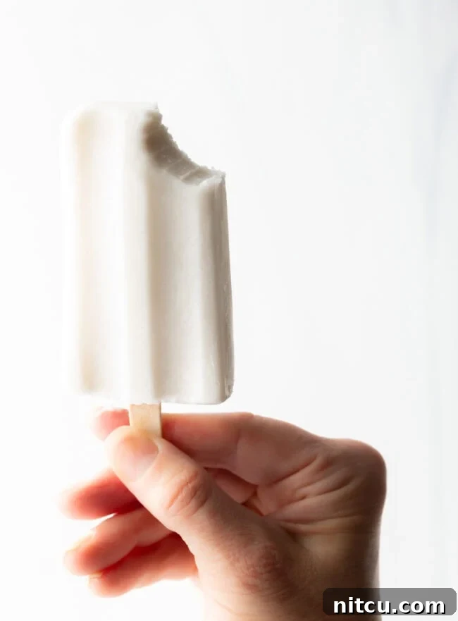 Creamy Dairy-Free Coconut Popsicles 11 A hand holding a coconut milk popsicle with a bite taken, revealing its creamy texture against a blurred background.