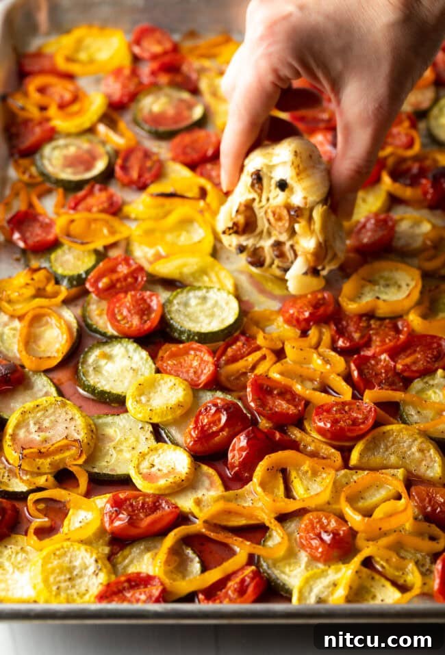 Creamy Burrata and Roasted Summer Harvest Pasta 9 Hand squeezing out soft roasted garlic cloves over the pan of roasted vegetables.