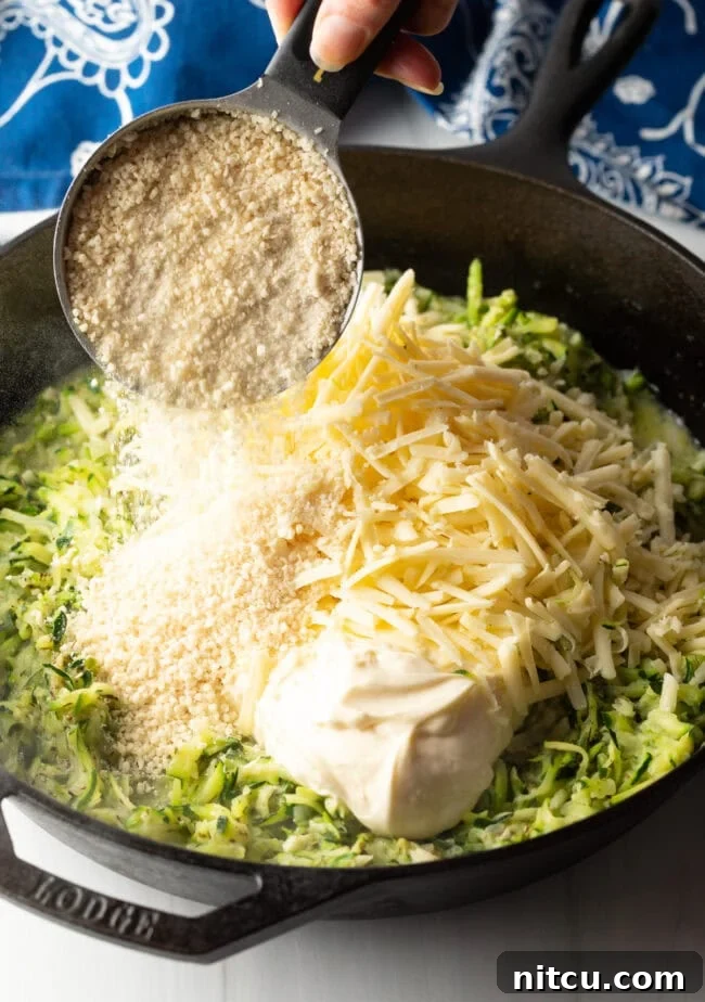 Golden Zucchini Gratin 7 Hand with measuring cup adding Panko breadcrumbs to the skillet with zucchini, mayo, and shredded cheese.