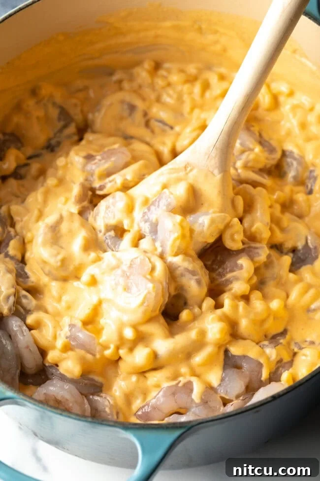 Decadent Shrimp Macaroni and Cheese 9 Wooden spoon stirring cheesy mac and cheese with uncooked shrimp in a large cooking pot.