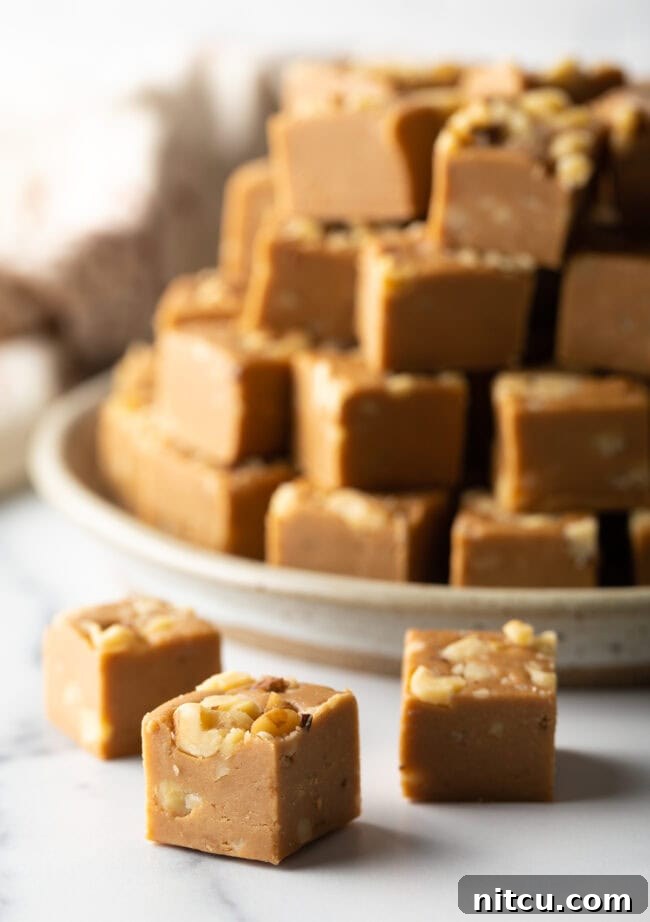 Creamy Maple Walnut Delight 2 Squares of maple walnut fudge in front of a stack of fudge, highlighting the beautiful texture and rich color.