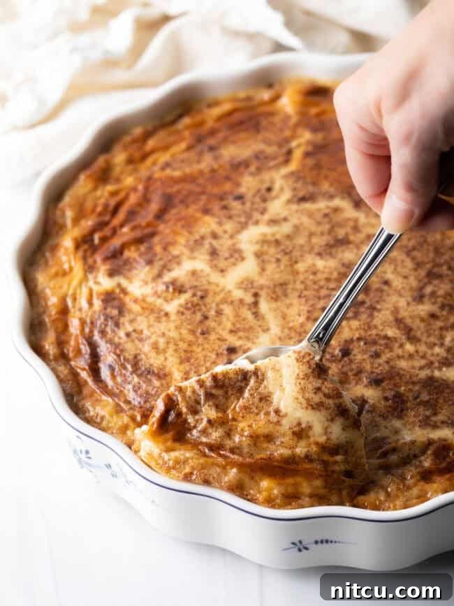 Golden Baked Rice Pudding 9 A hand scooping a generous serving of baked rice pudding from a large rectangular baking dish using a metal spoon.
