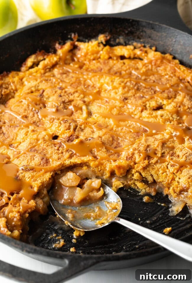 Gooey Caramel Apple Bake 12 A close-up view of the baked Caramel Apple Dump Cake in a cast-iron skillet, with a serving spoon removing a portion, highlighting its soft, gooey texture.