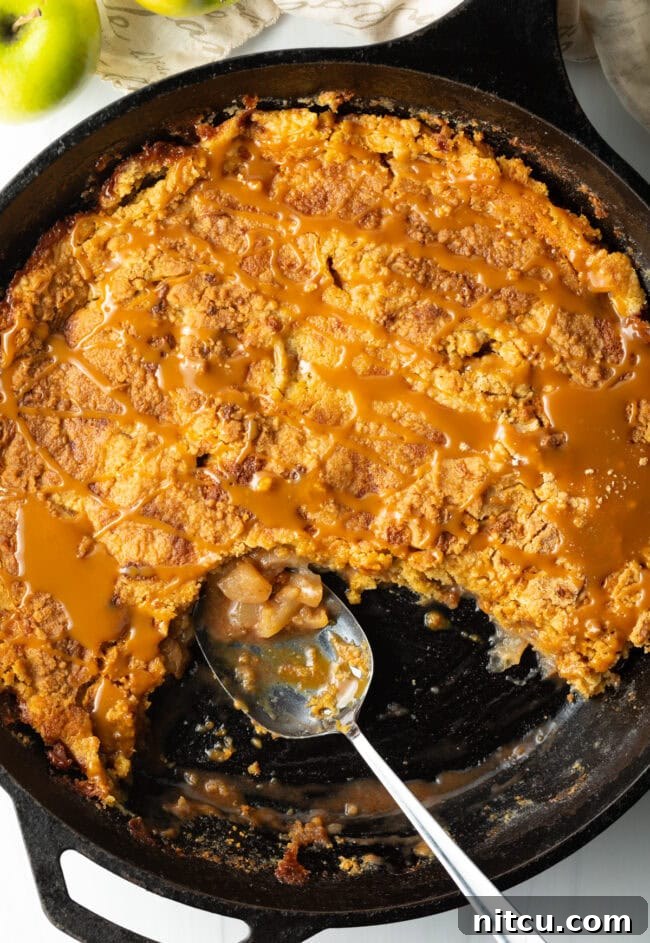 Gooey Caramel Apple Bake 3 A top-down view of a golden-brown Caramel Apple Dump Cake baked perfectly in a cast-iron skillet, with a portion already removed by a metal spoon, showcasing its gooey center.