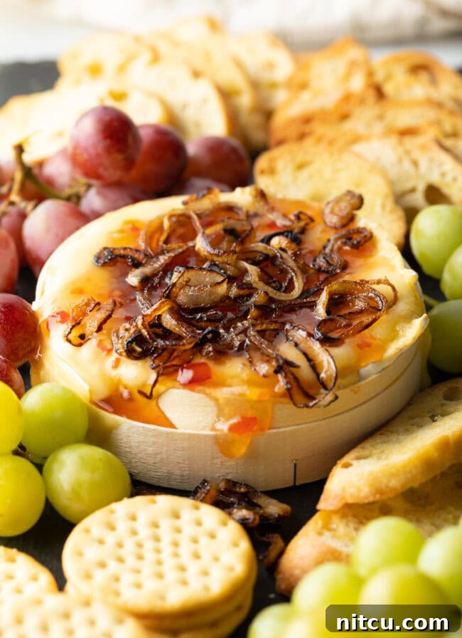 Gooey Baked Camembert Delight 2 Wheel of Camembert cheese baked with sweet chili sauce and sauteed shallots. It is on a platter and surrounded by red and green grapes, crackers, and baguette slices.